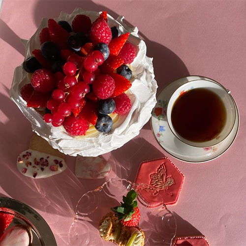Çiftehavuzlar'daki Art Cafe'de, pembe bir masanın üzerinde, çilek, ahududu, yaban mersini ve frenk üzümü gibi taze orman meyveleriyle süslenmiş büyük bir pavlova tatlısı. Tabağın yanında zarif bir fincanda çay ve farklı kurabiye çeşitleri de duruyor.