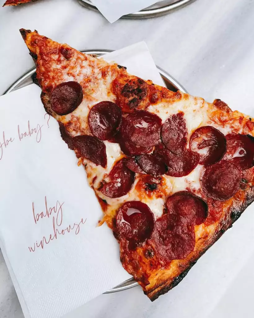 Beyaz bir peçete üzerinde servis edilen, üzeri kızarmış erimiş peynir ve çıtır sucuk dilimleriyle dolu iştah açıcı bir pizza dilimi, peçetenin sol tarafında kırmızı el yazısıyla yazılmış dikkat çekici baby winehouse markasıyla öne çıkıyor.