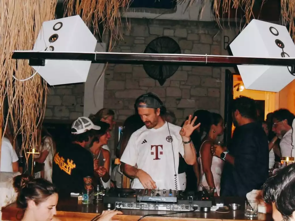 Bir barda, hasır tavan süslemelerinin ve altıgen hoparlörlerin altında, kulaklığında sakallı bir DJ, Pioneer mikseri başında kalabalık bir dinleyici kitlesine müzik çalarken bir elini havada tutarak performans sergiliyor, eğlenceli ve hareketli bir gece atmosferi sunuyor.