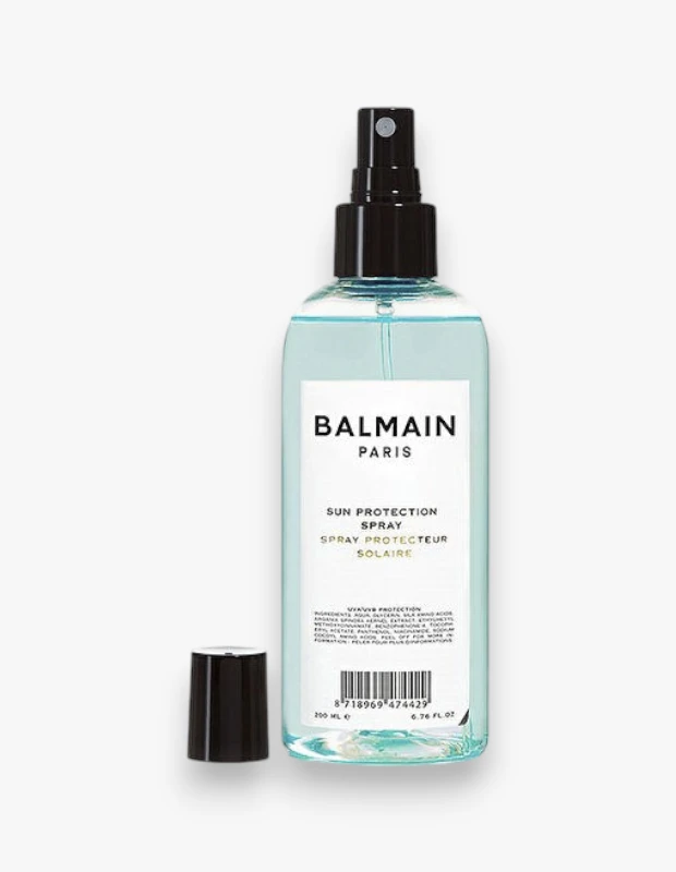 Beyaz arka plan üzerinde duran, Balmain Paris markalı, kapaksız ve siyah püskürtücü ağızlı, içinde açık mavi sıvı bulunan 200 ml'lik bir güneş koruyucu saç spreyi şişesi, ürünün koruyucu özelliklerini vurguluyor.