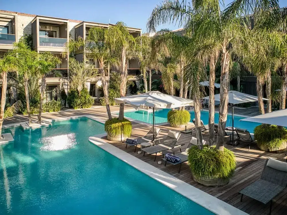 Biblos Resort Alaçatı'da, ışıl ışıl parlayan turkuaz renkli havuzların etrafında palmiye ağaçları, şemsiyeler ve şezlonglarla dolu ahşap bir güverte, modern mimariye sahip otel binalarının önünde huzurlu bir tatil ortamı sunuyor.
