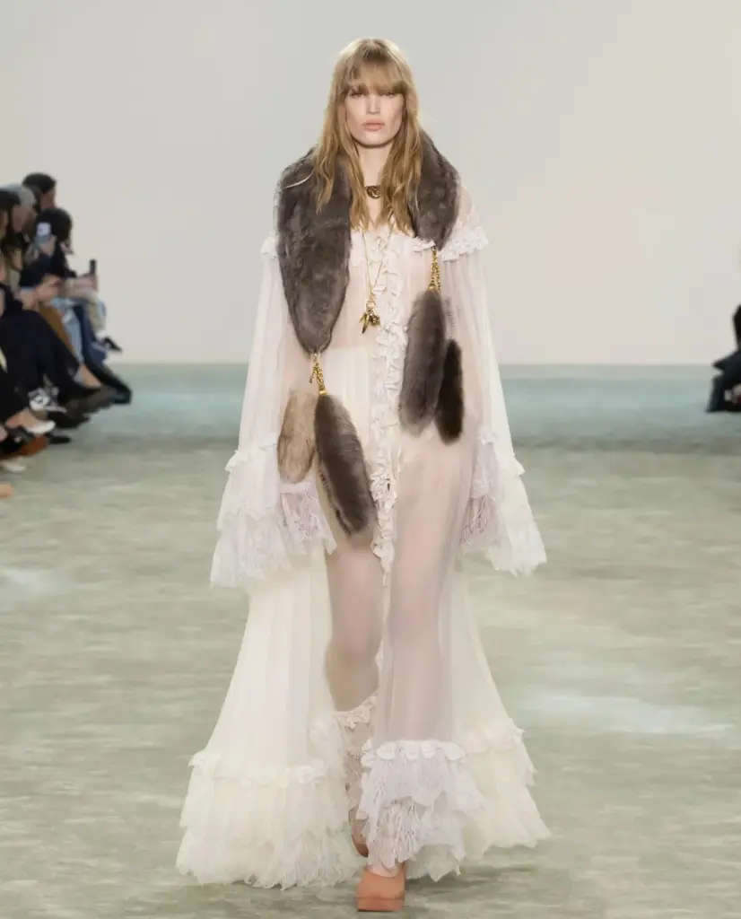 Beyaz dantel detaylı romantik maxi elbise ve omuza düşen kürk şal ile yürüyen model, bohem couture estetiğini modern yorumuyla sergiliyor.