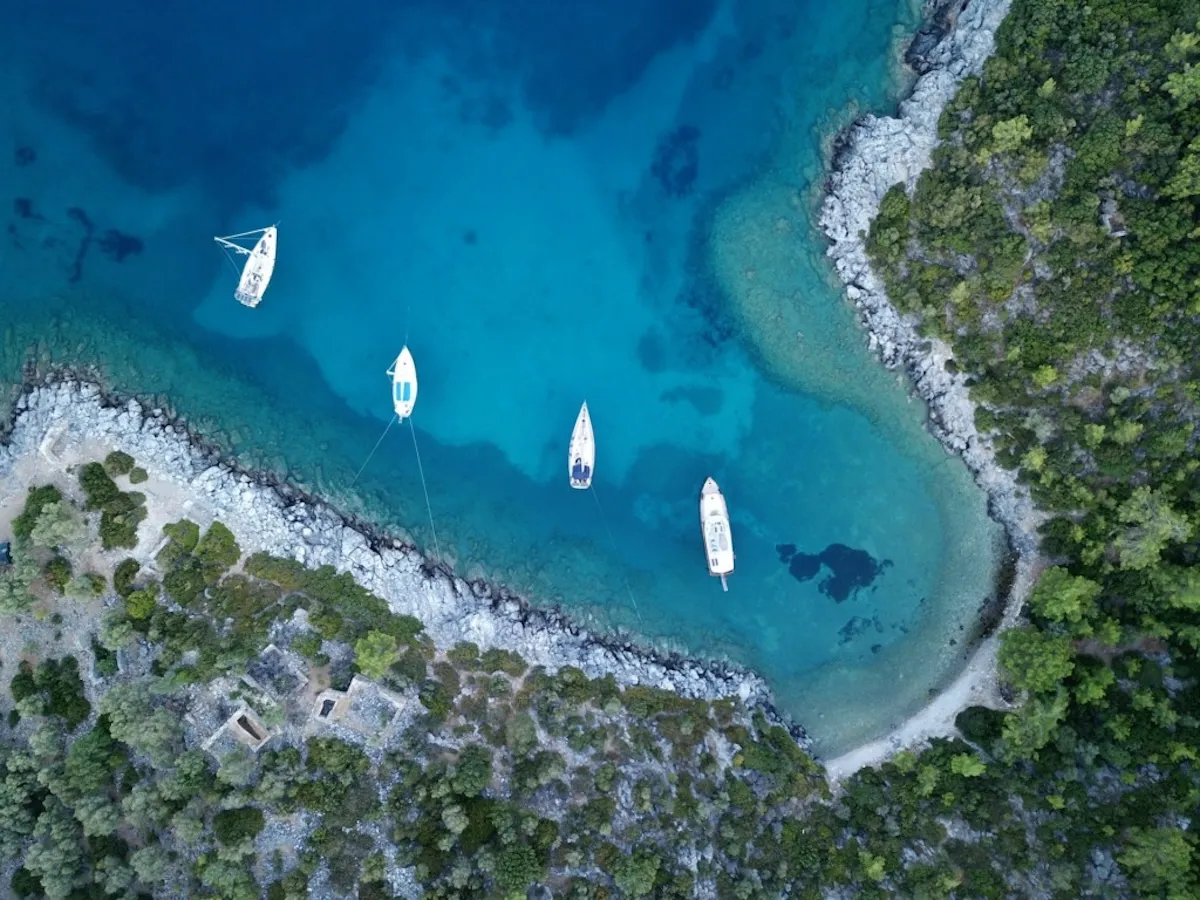 Bodrum seyahat rehberi: Bodrum'daki Cennet Koyu'nun havadan çekilmiş bu nefes kesici görüntüsünde, berrak turkuaz sularında dört beyaz tekne demirlemiş, etrafı yemyeşil ağaçlar ve kayalık kıyılarla çevrili doğal bir güzellik sergileniyor.