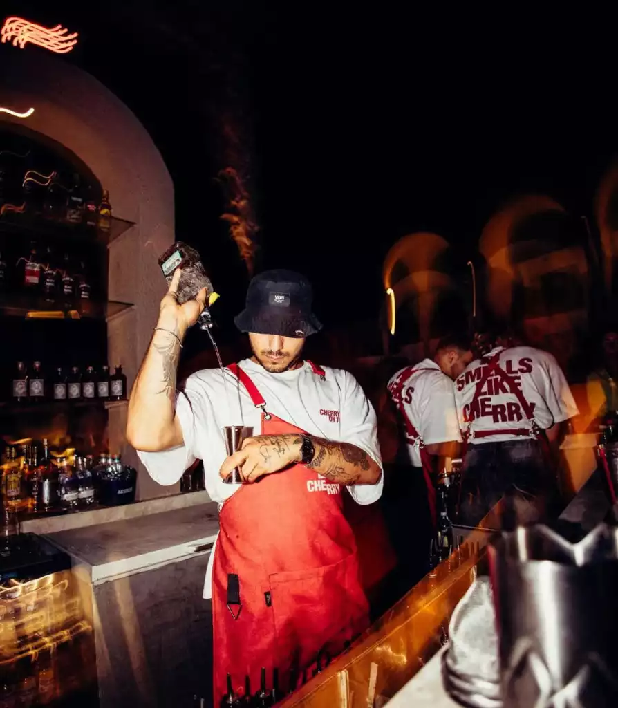 Karanlık bir bar tezgahının arkasında, siyah kova şapka ve kırmızı önlük giymiş, kolları dövmeli bir barmen, elindeki şişeden metal bir shakera özenle içecek dökerken görülüyor; üzerinde Cherry yazan tişörtü ve arkadaki Smells Like Cherry yazılı tişörtler, mekanın Cherry temasına vurgu yapıyor.