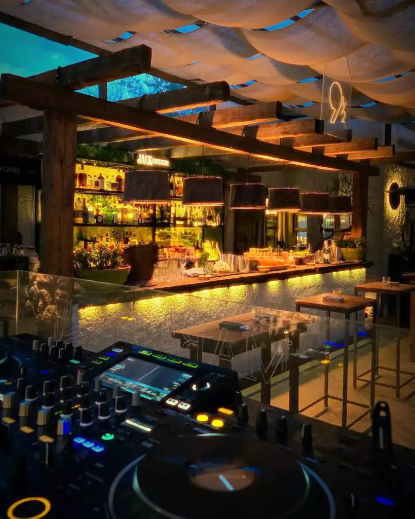 Ön planda renkli ışıklarla parlayan bir DJ mikseri ve pikap bulunurken, arka planda aydınlatılmış alkol şişeleriyle dolu uzun bir bar tezgahı ve ahşap tavanın altındaki şık Dokuzbucuk neon tabelası, mekânın eğlenceli ve modern atmosferini yansıtan canlı bir gece sahnesi sunuyor.