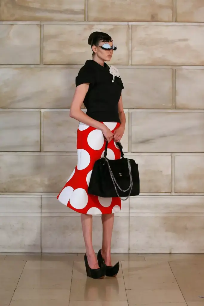 Parlak ışıkta duran model, belirgin kirpikli gözlükleri, modern siyah bluzu ve abartılı beyaz puantiye desenli kırmızı eteğiyle, Marc Jacobs'ın Minnie Mouse'tan ilham alan avangart puantiye trendini yansıtan cesur ve fütüristik bir moda yorumunu sergiliyor.
