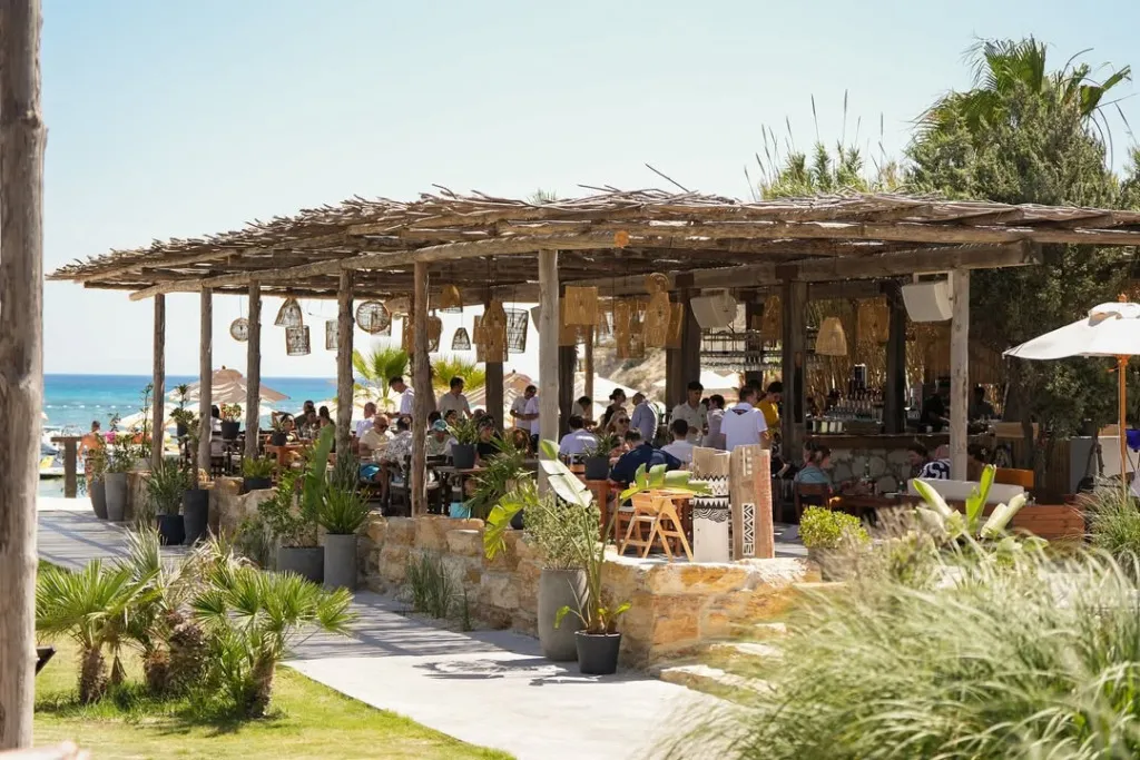 Güneşli bir havada, rustik ahşap pergolalı, denize sıfır Fly-Inn Beach adlı hareketli bir tesiste, hasır lambalar ve yeşil bitkilerle süslenmiş bar ve oturma alanlarında çok sayıda misafir yemek yiyip içeceklerin tadını çıkarırken, arka planda masmavi deniz uzanıyor.