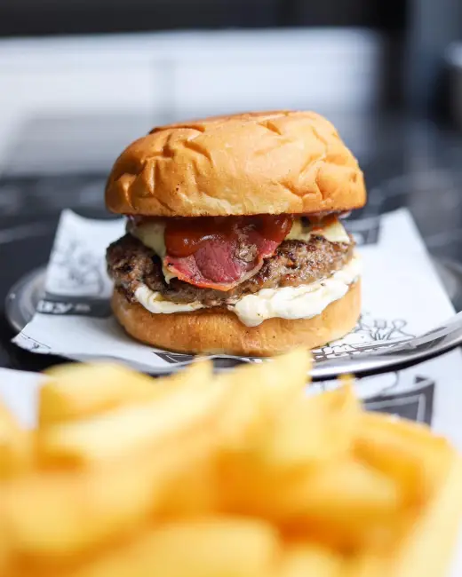 Şaşkınbakkal'da bulunan Grillin Guys restoranında, sunum tabağı içinde, üzerinde pastırma, peynir ve özel soslar bulunan bir hamburgerin yakın çekim fotoğrafı. Ön planda ise net olmayan bir şekilde patates kızartması görünüyor.