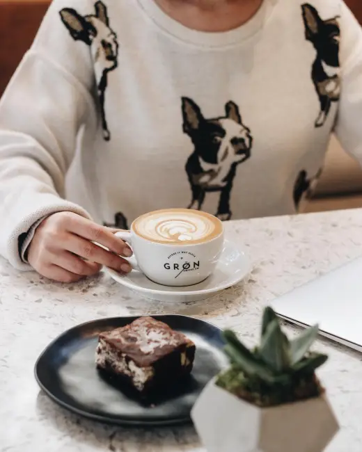 Caddebostan'daki Grön Coffee'de oturan bir kişinin, üzerinde köpek desenli bir kazakla kahve içtiği anın yakın çekim fotoğrafı. Masanın üzerinde, markanın logolu fincanında süt köpüklü kahve, küçük bir tabakta bir dilim brownie ve bir sukulent bitkisi görünüyor.