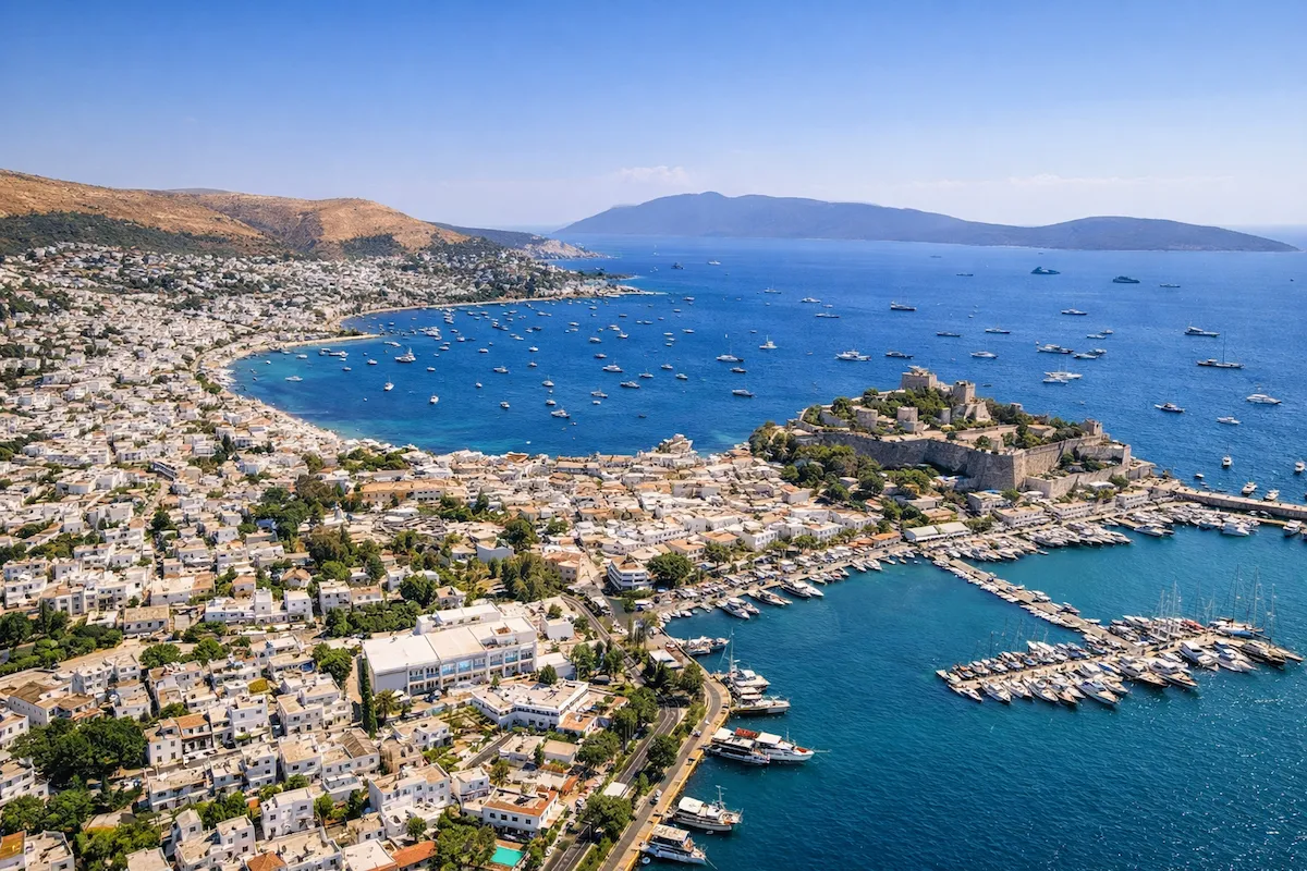 Bodrum seyahat rehberi: Gümbete yakın bir konumdan Bodrum'un masmavi körfezini, tarihi kalesini ve beyaz badanalı evleriyle uzanan sahil şeridini kuşbakışı gösteren bu panoramik fotoğrafta, denizde sayısız tekne ve yat hareketli bir yaz gününü yansıtırken, şehrin ikonik silüeti dikkat çekiyor.