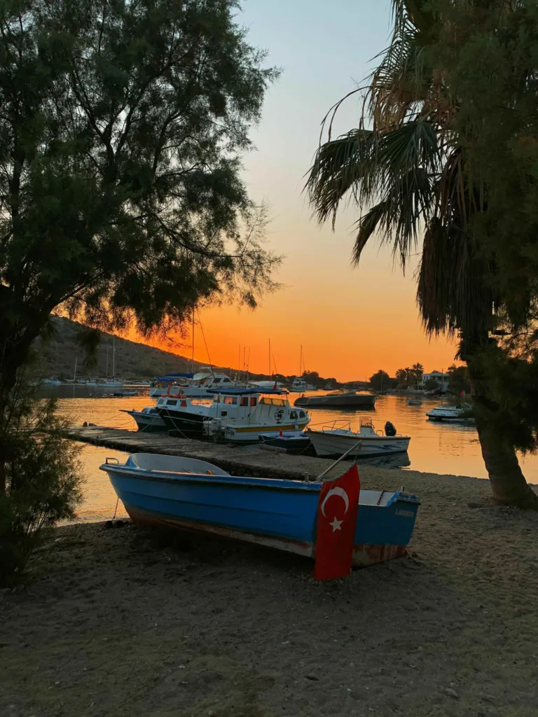 Bodrum seyahat rehberi: Gümüşlük, Bodrum'da gün batımının kızıl ve turuncu tonlarına bürünmüş gökyüzü altında, kumsala çekilmiş mavi renkli bir teknenin üzerinde asılı Türk bayrağı ve denizde demirli diğer teknelerin eşsiz huzur veren manzarası.