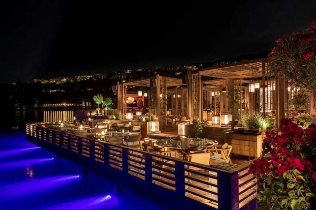 Gece çekilen bu büyüleyici geniş açılı fotoğraf, Bodrum Göltürkbükü'nde denizin üzerine kurulmuş, mavi ışıklarla aydınlatılmış sulara nazır lüks bir açık hava restoranı olan Hakkasan'ı gözler önüne seriyor; ahşap pergolalar, asılı fenerler ve zarif kırmızı çiçeklerle bezeli yemek alanları, uzaktaki ışıklarla birlikte sıcak ve sofistike bir atmosfer yaratıyor.