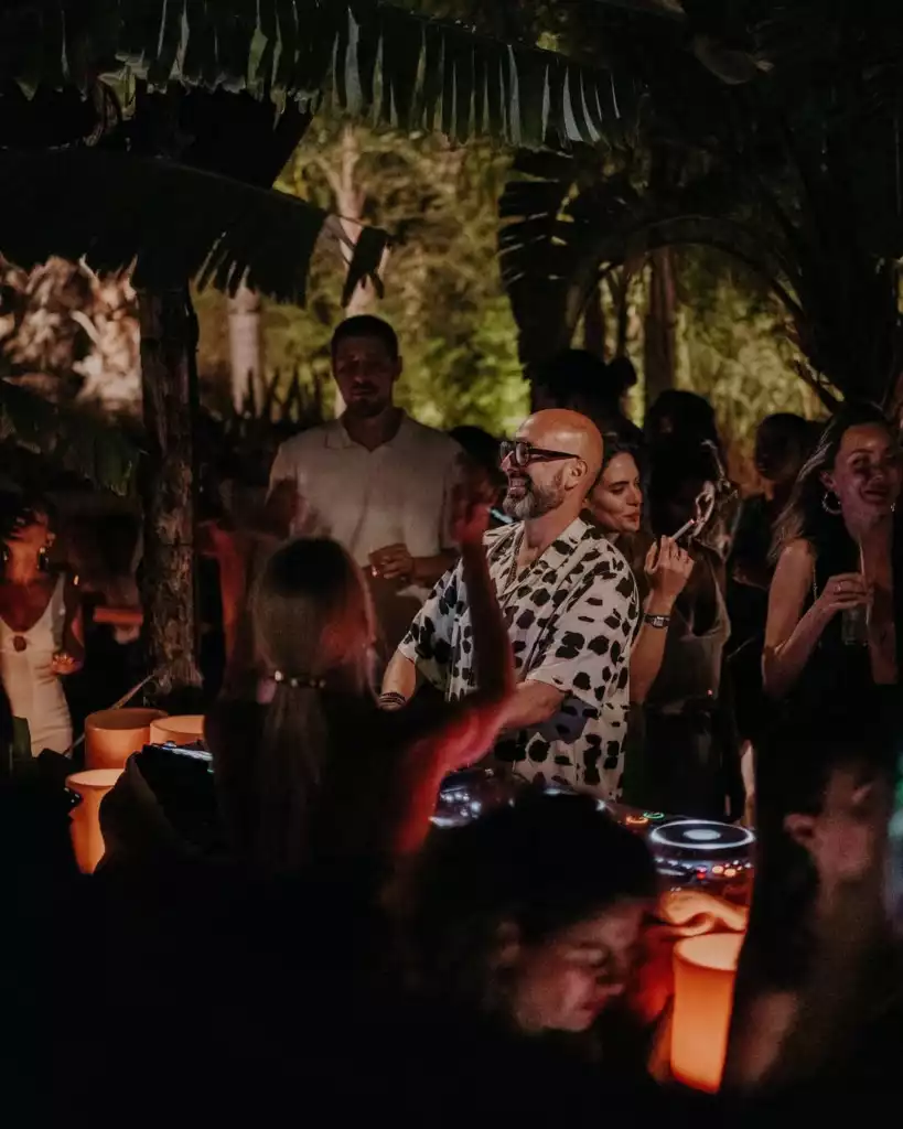 Kel, gözlüklü ve sakallı bir adamın desenli gömleğiyle gülümseyerek kalabalık bir açık hava gece partisinde DJ performansı sergilediği, etrafında dans eden insanların ve tropikal bitki örtüsü arasında sıcak ambiyans ışıklarının aydınlattığı canlı bir sahne.