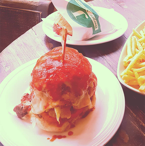 Caddebostan'daki J Burger restoranında masada servis edilen, üzerinde bol salçalı bir sos bulunan ıslak hamburger ve yanında bir tabak patates kızartması. Arka planda ambalajlı başka bir hamburger daha görünüyor. 