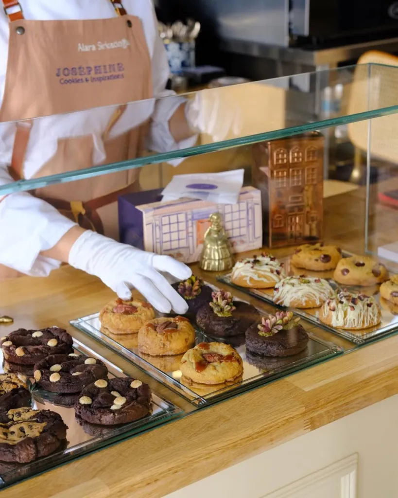 Joséphine Cookies'de, cam vitrinin arkasında, farklı lezzetlerdeki kurabiyelerin sergilendiği bir bölüm. Çikolatalı, bademli ve damla çikolatalı kurabiyeler ayna tepsiler üzerinde duruyor. Bir çalışan beyaz eldiveniyle bir kurabiyeyi yerleştiriyor.