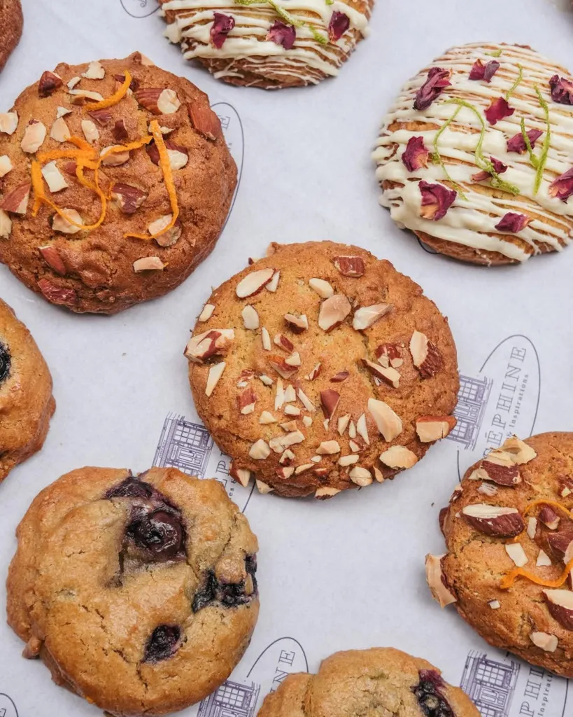 Joséphine Cookies'in farklı lezzetlerdeki kurabiyelerinin yakın çekim fotoğrafı. Kurabiyelerden bazılarında badem ve portakal kabuğu, bazılarında beyaz çikolata sosu ve gül yaprakları, bazılarında ise yaban mersini parçacıkları görülüyor.