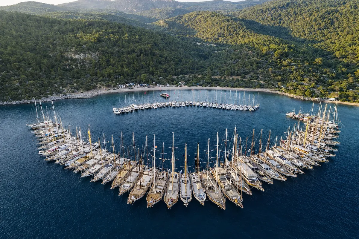 Bodrum seyahat rehberi: Mazikoy yakınlarındaki Kissebuku Koyu'nun yemyeşil tepelerle çevrili berrak turkuaz sularında, büyük bir hilal formasyonu oluşturmuş onlarca zarif yelkenli ve geleneksel gulet teknesi, huzurlu bir akşamüstü veya sabah manzarasını sergiliyor.