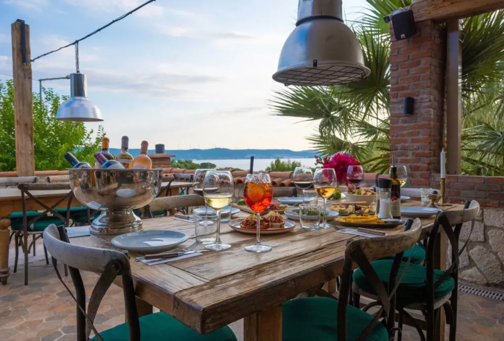 Deniz manzaralı bir açık hava restoranında, rustik ahşap masa üzerinde çeşitli şaraplar, turuncu bir kokteyl ve lezzetli yemeklerle donatılmış, yeşil minderli sandalyelerle çevrili keyifli bir yemek deneyimi sunuluyor.