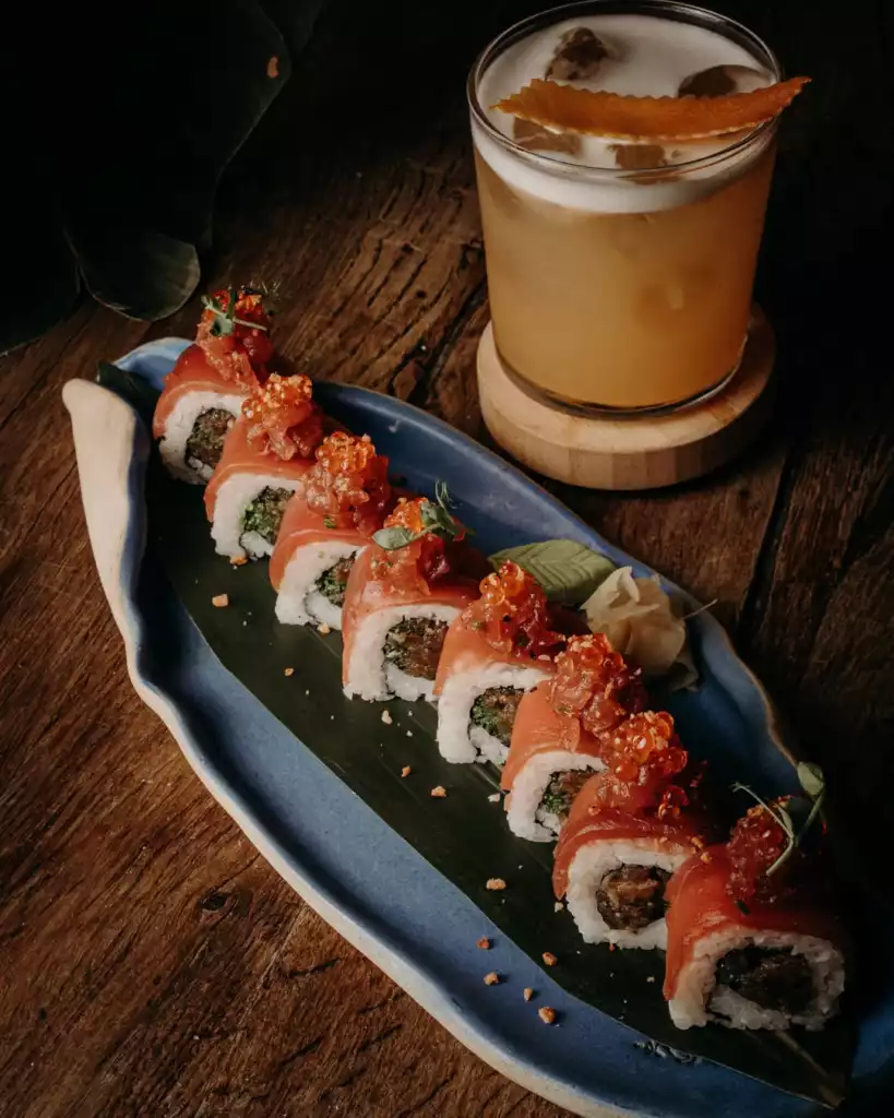 Ahşap bir masa üzerinde, mavi, yaprak formunda şık bir tabakta sunulan, üzerine parlak turuncu ikura ve yeşilliklerle bezenmiş, somon dilimleriyle sarılı lezzetli sushi ruloları ve yanında ferahlatıcı köpüklü bir kokteylin cazip bir görseli.