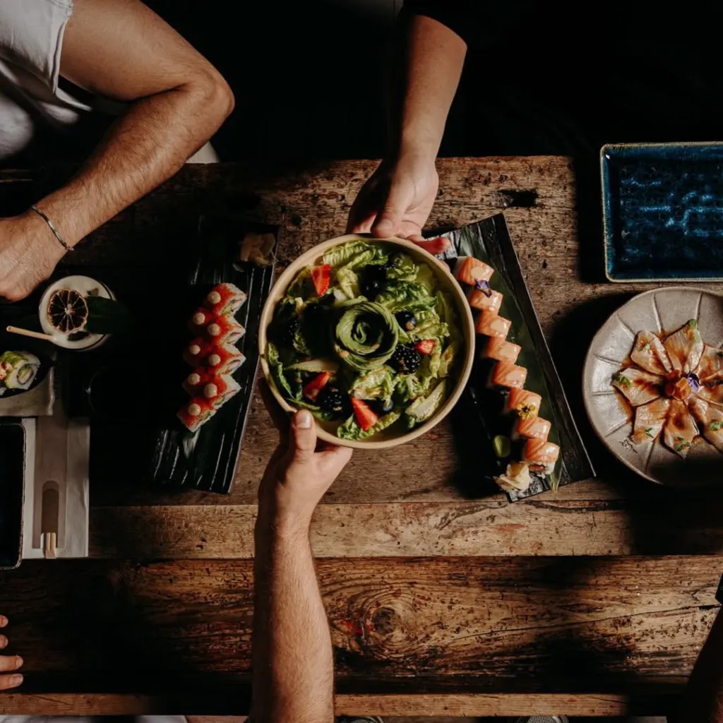 Birden fazla elin bir masa etrafında, avokado, çilek ve böğürtlenle süslenmiş taze yeşil salata, somonlu suşi rolları, üzeri kırmızı toplu suşi ve çiçek şeklinde düzenlenmiş sashimi gibi özenle hazırlanmış çeşitli yemekleri paylaştığı şık bir Alaçatı restoran deneyimi.