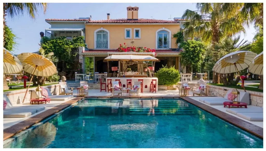 La Capria Suite Hotel'in huzur dolu dış mekanında, berrak turkuaz renkli yüzme havuzunun etrafında, konforlu şezlonglar, dekoratif kırmızı yan masalar ve zarif sarı-beyaz şemsiyelerle donatılmış, arkada ise çiçeklerle bezenmiş çekici bir havuz barının yer aldığı Akdeniz esintili lüks bir konaklama ortamı görünüyor.