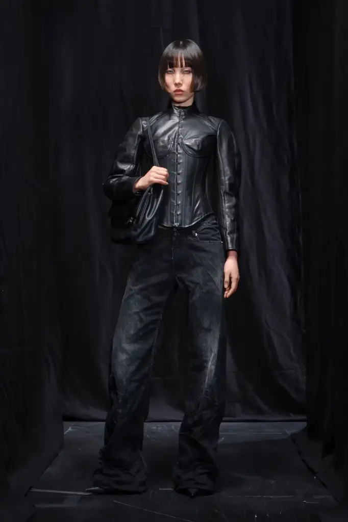 Siyah deri korsaj detaylı fitted ceket ve geniş paça koyu denim pantolon ile poz veren model, 2025 koleksiyonlarının neo-grunge ve fetiş estetiğini birleştiren güçlü bir görünüm sunuyor.