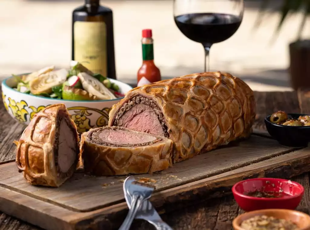 Ortasından kesilmiş, pembe ve sulu etli, üzeri ızgara desenli altın rengi çıtır milföy hamuruyla kaplı iştah açıcı bir Beef Wellington, rustik ahşap bir kesme tahtası üzerinde sunuluyor; arka planda salata, kırmızı şarap kadehi, zeytinler ve sos şişeleri görülerek zengin bir öğün tablosu oluşturuyor.
