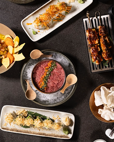 Suadiye'de bulunan Machi restoranında koyu renkli bir masa üzerinde, farklı Japon ve Peru yemeklerinin yer aldığı bir sofra. Masada sashimi, şişler, patates cipsi, karides krakerleri ve çeşitli soslar görülüyor.