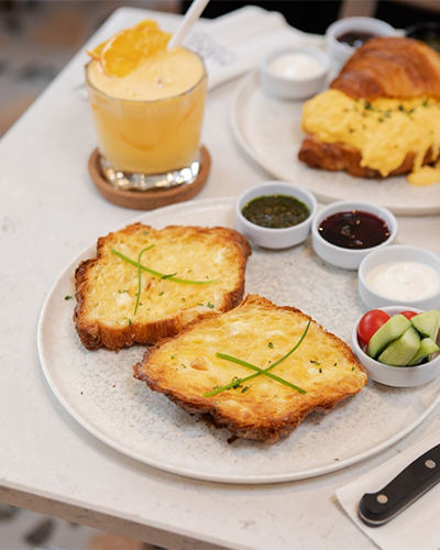 Manu Bakery & Eatery'de servis edilen, masanın üzerinde peynir, maydanoz ve otlar ile süslenmiş bir dilim ekmek. Tabağın yanında yeşil sos, reçel, yoğurt ve salatalık-domates dilimleri bulunuyor. Arka planda ise kruvasan ve meyve suyu görünüyor.