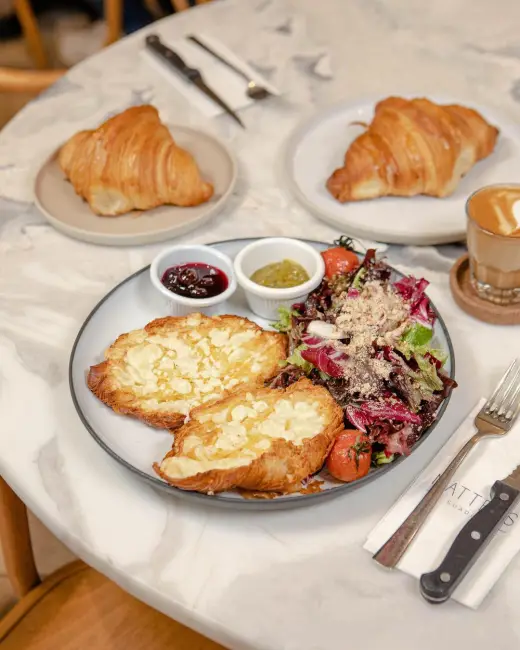 Suadiye'deki Matters restoranında masada servis edilen bir kahvaltı tabağı. Tabakta salata ve yanında reçel ve pesto sos gibi iki sosluk bulunuyor. Ana tabağın solunda bademli ve peynirli iki adet hamur işi, sağında ise marul, domates ve kırmızı soğanlı bir salata yer alıyor. Masada ayrıca iki adet sade kruvasan, çatal bıçak takımı ve bir bardak kahve de görülüyor.