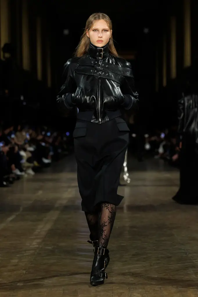 Siyah yüksek yakalı deri ceket, midi etek ve dantel çoraplarla podyumda yürüyen model, keskin hatlı modern gotik lüksü temsil ediyor.