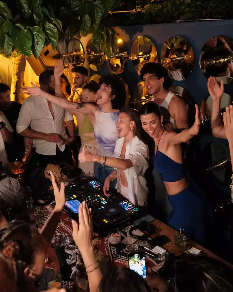 Bir gece kulübünde veya açık hava barda, bir DJ’in mixer başında coşkuyla müzik çaldığı, çevresindeki kalabalığın ise kolları havada dans edip neşeyle şarkı söylediği, genel olarak enerjik ve eğlence dolu bir parti atmosferi sunuluyor; bu an, gençlerin bir araya gelerek müziğin tadını çıkardığı hareketli bir kutlamayı gözler önüne seriyor.