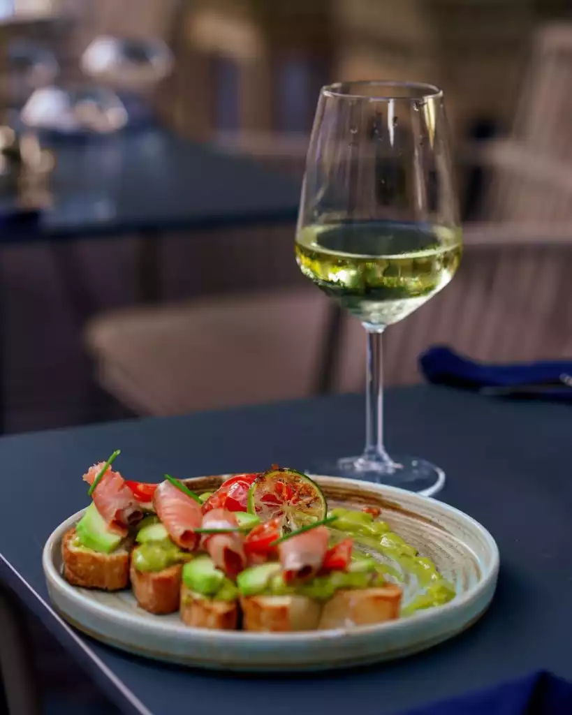 Bir restoranda koyu renk bir masanın üzerinde, taze avokado ezmeli, somon fümeli, çeri domatesli ve frenk soğanlı lezzetli bruschetta benzeri atıştırmalıkların sunulduğu bir tabak ile yanında sarımsı beyaz şarap dolu şık bir kadeh göz alıcı bir şekilde duruyor.