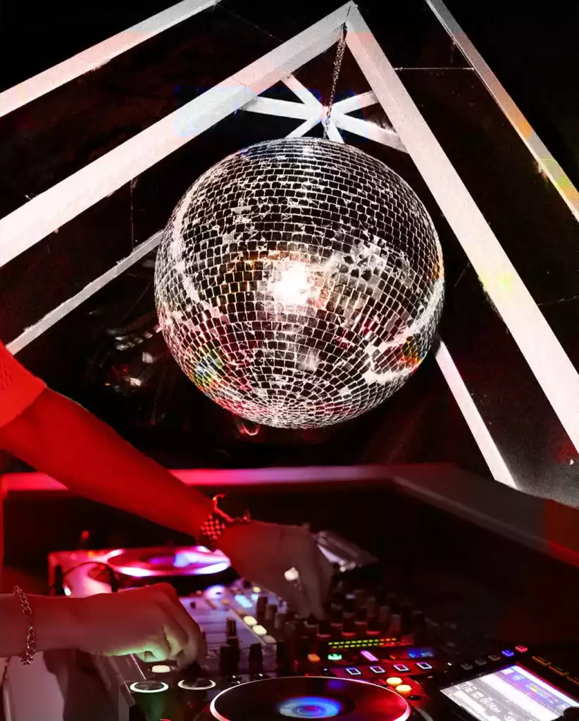 Loş ve kırmızı tonlardaki bir gece kulübü ortamında, tavandan sarkan büyük, aynalı bir disko topu parlarken, bir DJ'in elleri müzik mikseri üzerindeki ışıklı tuşları ve döner plakları ustaca ayarlayarak coşkulu bir atmosfer yaratıyor.