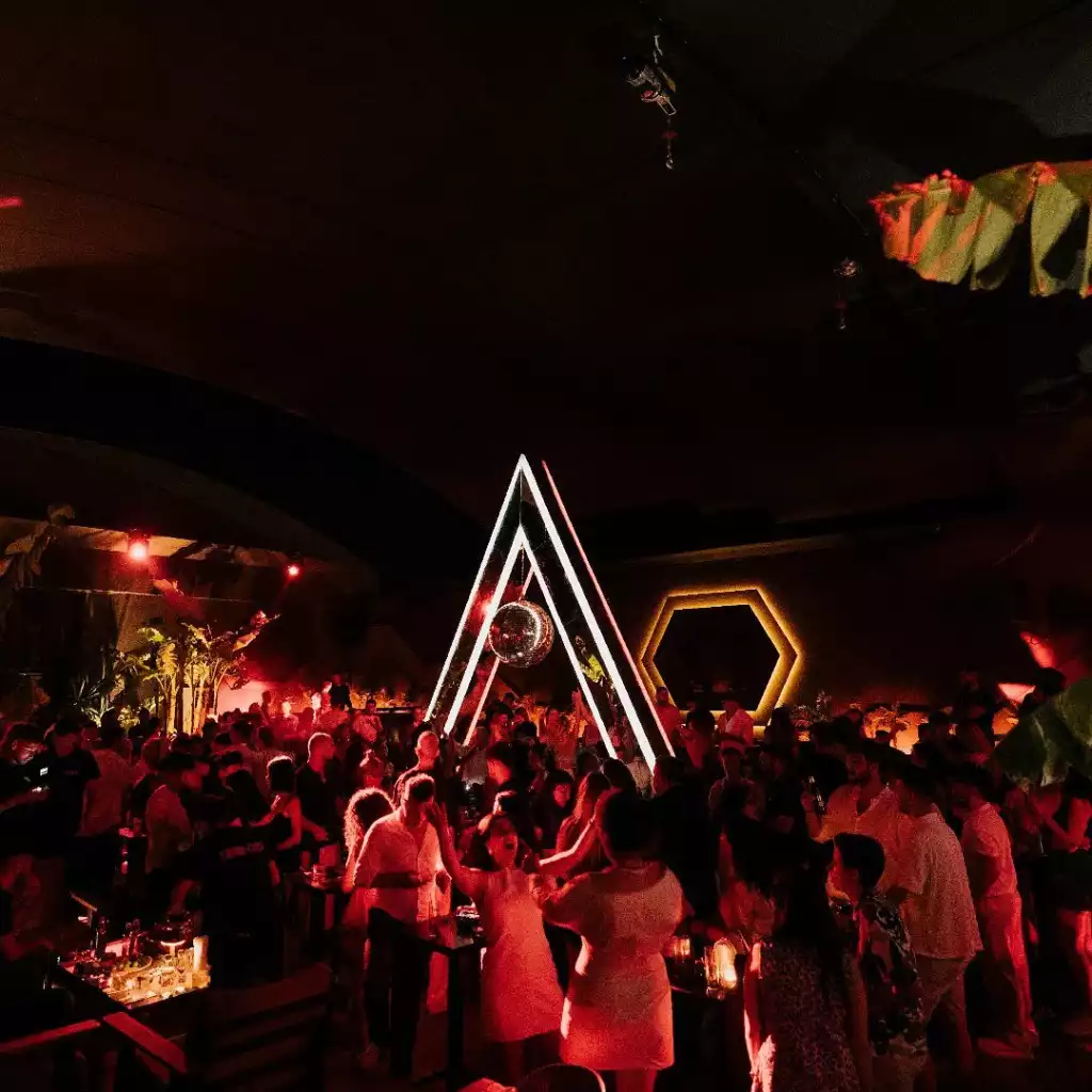 Kırmızı tonlarda aydınlatılmış, büyük bir disko topu ve neon ışıklı üçgen kemerle süslenmiş bir gece kulübünde, müziğin ritmine uygun şekilde dans eden ve sosyalleşen kalabalık bir grup insan görülüyor.
