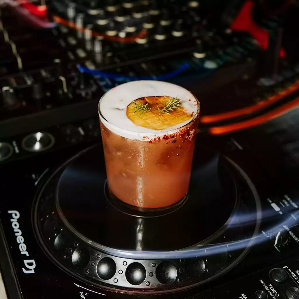Pioneer DJ pikapının üzerinde duran, kenarı baharatla kaplı, üzerinde kalın beyaz köpük, ızgara ananas dilimi ve biberiye dalı olan bu özel kokteyl, gece kulübü ambiyansının ve müziğin keyfini çıkarmanız için sizi bekliyor.