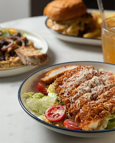 Suadiye'deki Nove Garden'da masada servis edilen, üzerinde rendelenmiş parmesan peyniri olan ve domates, marul ve mısırla hazırlanan tavuklu sezar salata. Arka planda hamburger ve patates kızartması da görünüyor.
