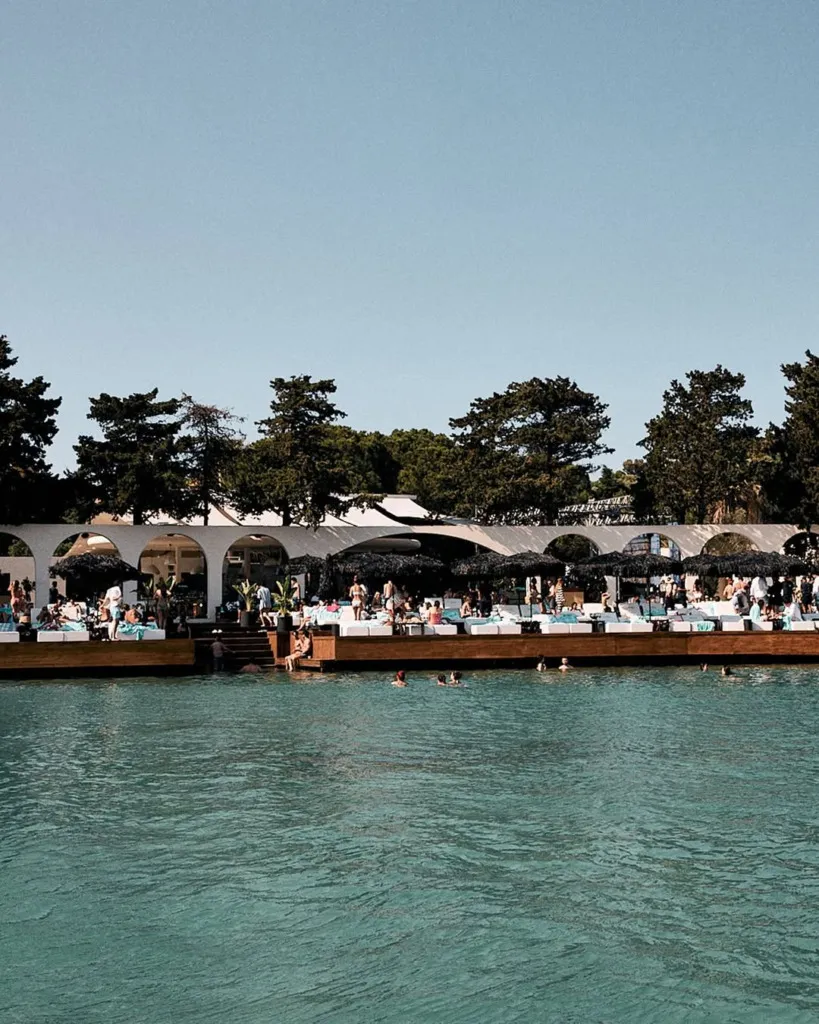 Parlak güneşli bir günde, turkuaz renkteki suyun kenarında konumlanan Om Paparazzi Beach Club'da, beyaz kemerli yapının önündeki ahşap platformda şemsiyelerin altında dinlenen ve denizde yüzen birçok kişi keyifli anlar yaşıyor.