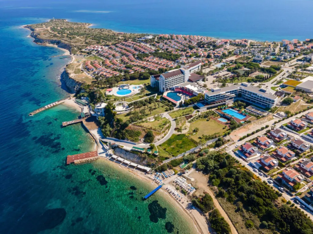 Çeşme'deki Onlive Deluxe Otel'in havadan çekilmiş görüntüsü, turkuaz renkteki Ege Denizi kıyısında konumlanmış, birden fazla yüzme havuzu, yemyeşil peyzajlı bahçeler, denize uzanan ahşap iskeleler ve çevresindeki konutlarla birlikte geniş bir tatil kompleksini gözler önüne seriyor.