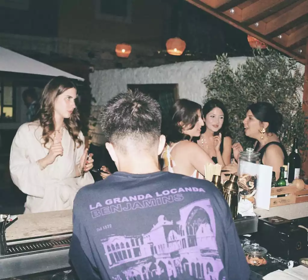 Gece bir açık hava barında geçen sahnede, üzerinde Venedik mimarisi deseni olan koyu renk tişörtlü, sırtı dönük bir kişinin baktığı yönde, bar tezgahının solunda telefon tutan uzun saçlı bir kadın ve sağında içeceklerin önünde sohbet eden üç kadın bulunuyor; arka planı ise ağaçlar ve sıcak ışıklı fenerler süslüyor.