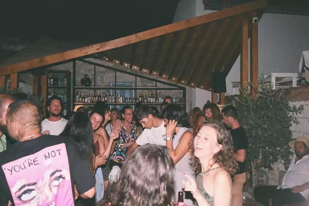 Gece çekilmiş bu karede, bir açık bar etkinliğinde kalabalık bir grup genç neşeli bir şekilde sohbet edip dans ediyor, arka planda ise çeşitli içkilerin sergilendiği bar rafları dikkat çekiyor.
