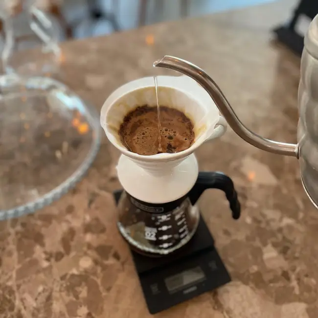 Caddebostan'daki Orbis Cafe'de, bir baristanın özel bir su ısıtıcısı kullanarak V60 demleme yöntemi ile kahve hazırladığı anın yakın çekimi. Fotoğrafta kahve filtreden geçerek altındaki sürahiye damlıyor ve hassas bir terazi üzerinde duruyor.