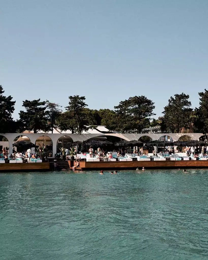 Güneşli bir günde, berrak turkuaz denizin kıyısında yer alan, beyaz kemerli modern bir yapının önünde sıralanmış koyu renk şemsiyelerin altında beyaz şezlonglarda keyif yapan ve denizde yüzen çok sayıda insanla dolu, gözde bir plaj kulübü manzarası.