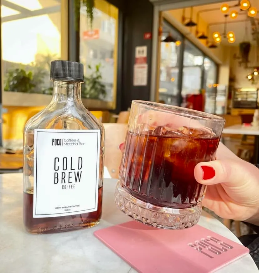 Suadiye'deki Poco Matcha'nın dışarıdaki masasında, bir elin tuttuğu buzlu cold brew kahve dolu bardak ve yanında şişe içinde markanın soğuk kahvesi görülüyor.