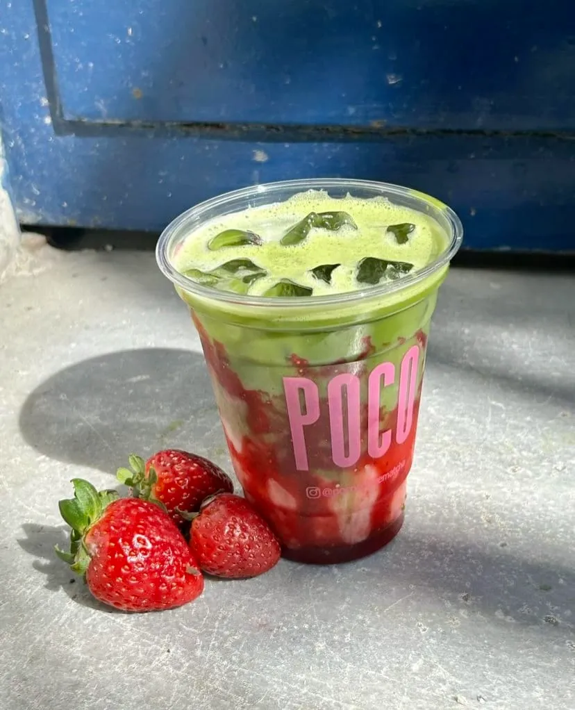 Poco Matcha'da sunulan, buzlu ve çilek soslu matcha içeceğinin yakın çekim fotoğrafı. İçecek, Poco logosunun bulunduğu şeffaf bir bardakta servis ediliyor ve yanında üç adet taze çilek bulunuyor.