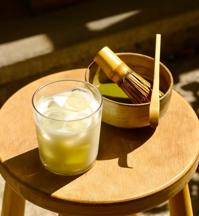 Poco Matcha'da ahşap bir tabure üzerinde buzlu bir matcha içeceği ve yanında matcha hazırlamakta kullanılan bambu kaşık ve çırpıcı gibi gereçlerin bulunduğu bir kase.
