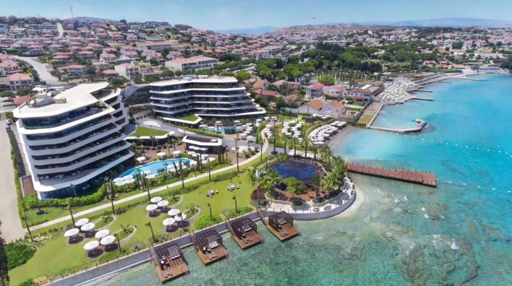 Çeşme'deki Reges Luxury Collection lüks otel ve spa tesisinin havadan görünümü; modern mimarili kıvrımlı binalarını, yemyeşil bahçelerdeki havuz ve şezlong alanlarını, berrak turkuaz denize uzanan iskeleleri ve su üstü özel dinlenme platformlarını, arka planda ise evleri sıralı tepeleri sergiliyor.