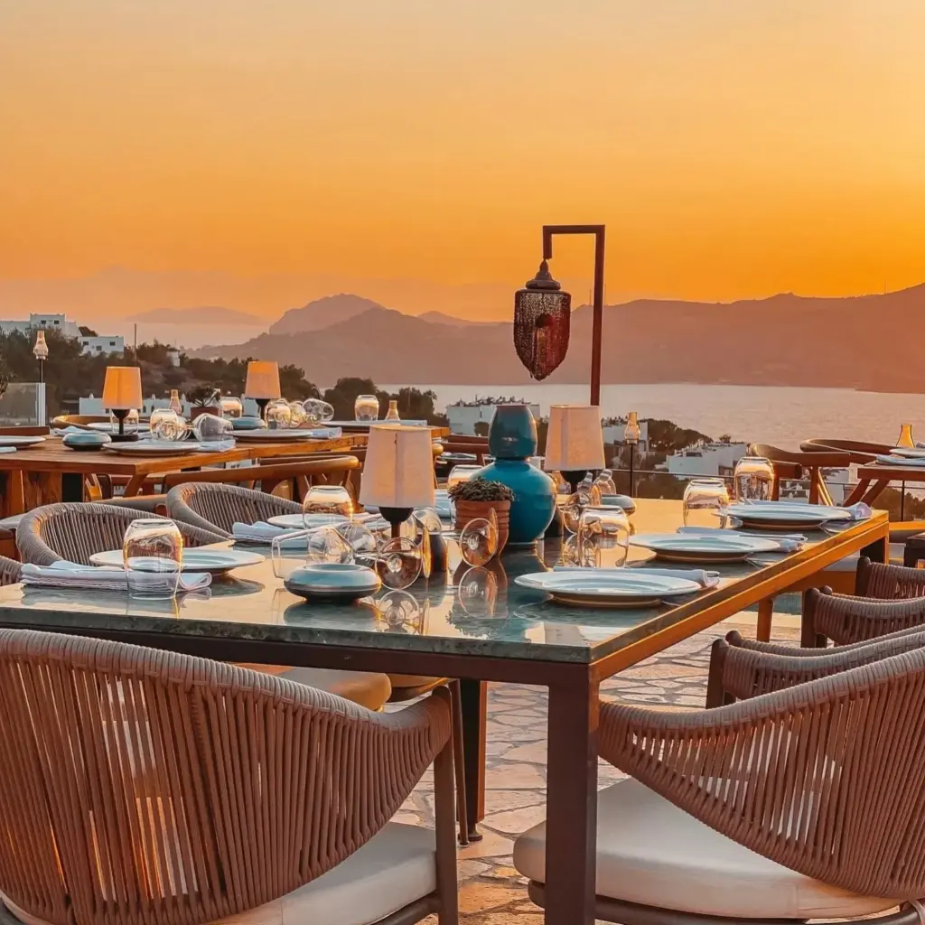 Yalıkavak'ta Ruby Mare adlı restoranın denize nazır terasında, turuncuya bürünmüş gün batımının nefes kesen manzarası eşliğinde, şık bir akşam yemeği için özenle dizilmiş masalar, hasır detaylı sandalyeler ve masalardaki dekoratif aydınlatmalar büyüleyici bir atmosfer sunuyor.