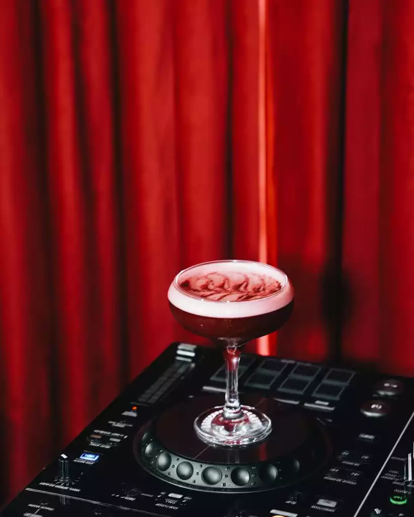 Siyah bir DJ mikseri üzerinde konumlandırılmış şık bir kadehte, üstünde beyaz köpük ve içinden beliren kırmızı desenler barındıran derin kırmızı bir kokteyl içeceği, arka plandaki dalgalı kırmızı sahne perdesiyle gece hayatının sofistike ve enerjik atmosferini vurguluyor.