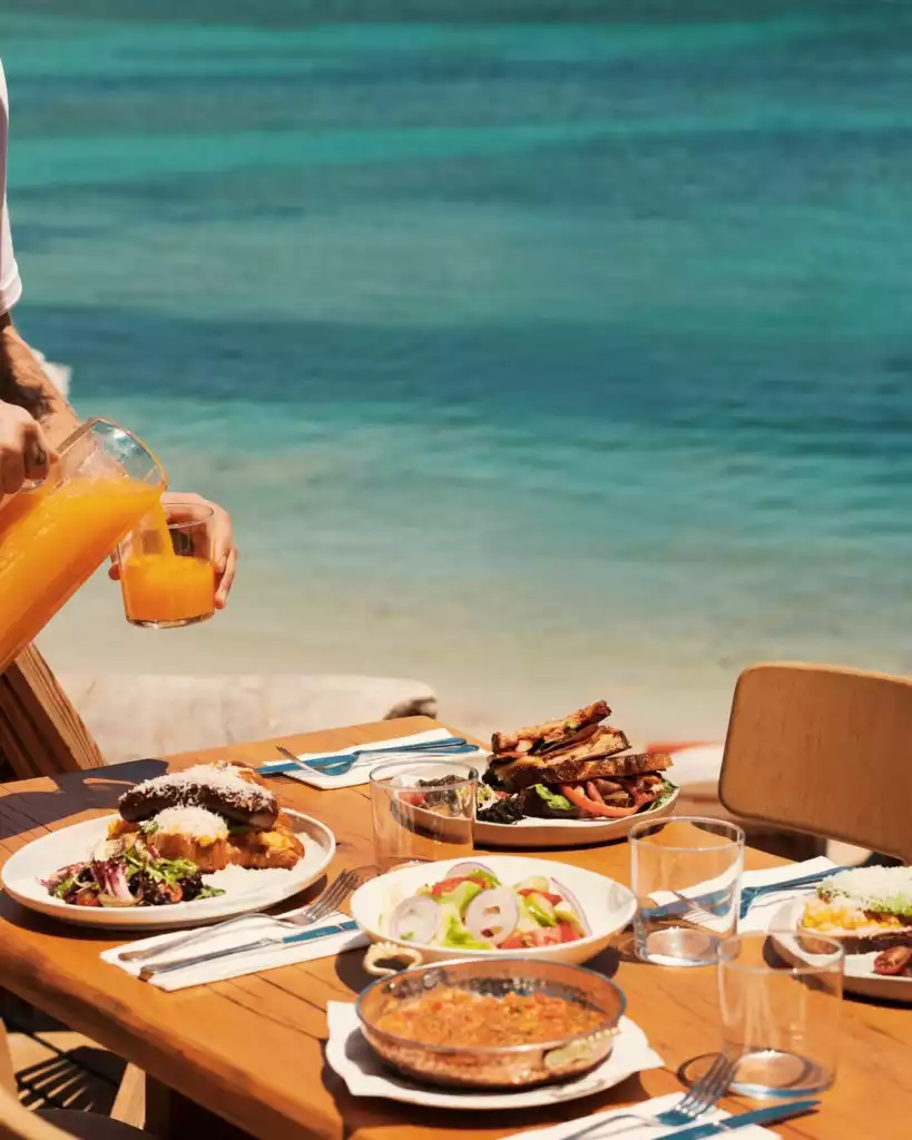 Türkiz denizin huzur veren manzarası eşliğinde, güneşli bir günde açık havada kurulan ahşap bir masada zengin bir kahvaltı veya brunch sofrası; bir elin sürahiden taze sıkılmış portakal suyunu bardağa doldurduğu bu an, menemen, kruvasan, sandviç ve salata gibi çeşitli lezzetleri gözler önüne seriyor.